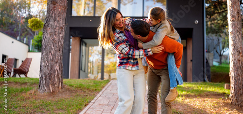Cheerful family with father carrying daughter piggyback, laughing and having fun outdoors on a sunny autumn day, enjoying bonding moments in front of their modern house