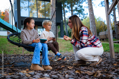 Mother squatting and engaging in conversation with her young daughter and son, who are sitting together on a outdoor swing and holding smartphones, enjoying time in the autumn backyard