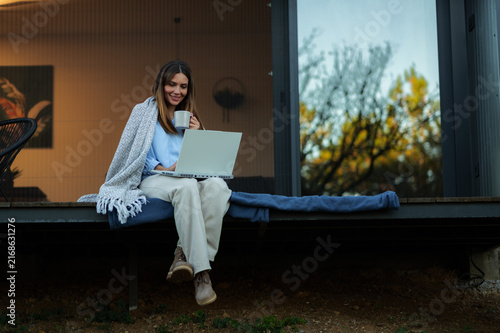 Young woman comfortably sitting on a modern terrace with a warm blanket, using a laptop and drinking from a cup, enjoying remote work and relaxation outdoors during autumn