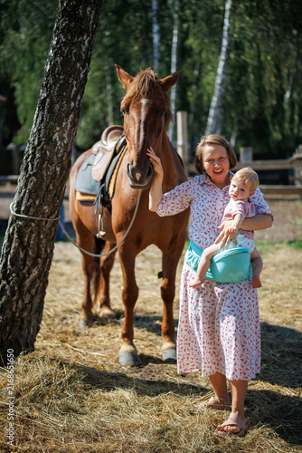 Woman holding a baby stands near a saddled brown horse outdoors in summer