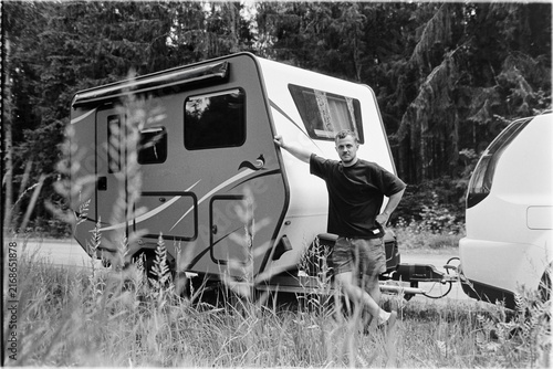 Black and white film photo of a man leaning against a caravan near a forest. The image exhibits visible grain