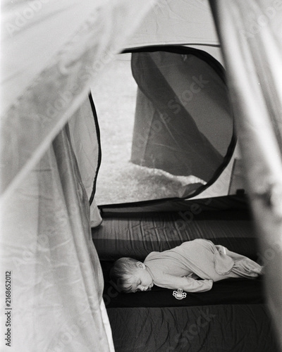 Monochrome film photograph of a baby sleeping peacefully on a mat inside a camping tent, showing characteristic film grain