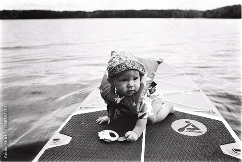 Low-angle shot on textured black and white film of a baby in a vest crawling on a paddle board on the water