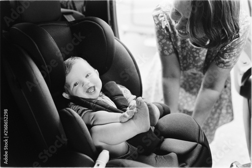 Candid analog film capture in black and white showing a smiling baby in a car seat with a woman looking on