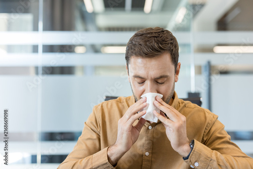 Konstfotografi Man in casual brown shirt blows his nose into a white tissue, looking unwell at