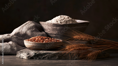 Artistic still life of whole rye grains and flour in ceramic bowls on natural linen fabric, scattered kernels and wheat ears, a rustic harvest composition for bakery and agriculture themes.