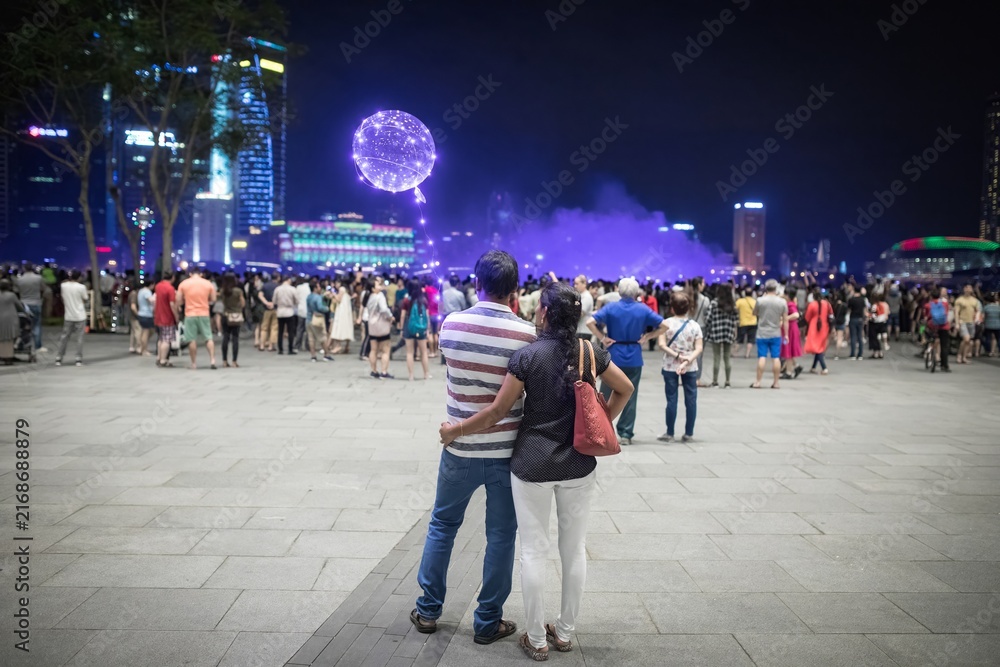 Obraz premium Couple stands embracing among crowd watching illuminated night light show with glowing sphere lantern and city skyline reflected in purple haze at Singapore waterfront promenade