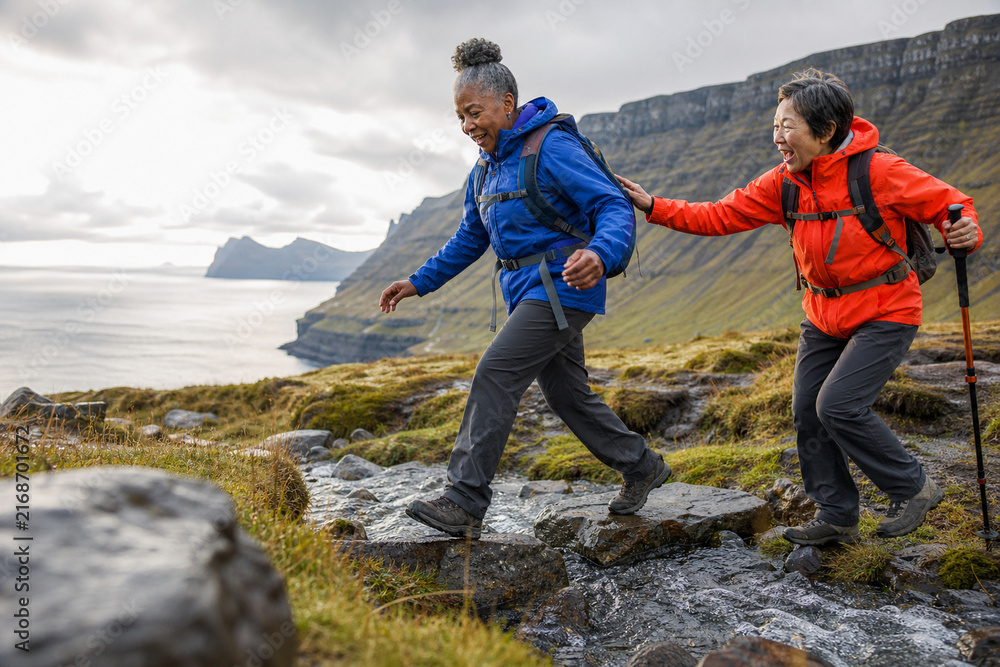 Fototapeta premium Two hikers in bright jackets traverse a rocky trail along a rugged coastline, supporting each other as they cross a shallow stream. A moment of teamwork, resilience, and outdoor adventure.