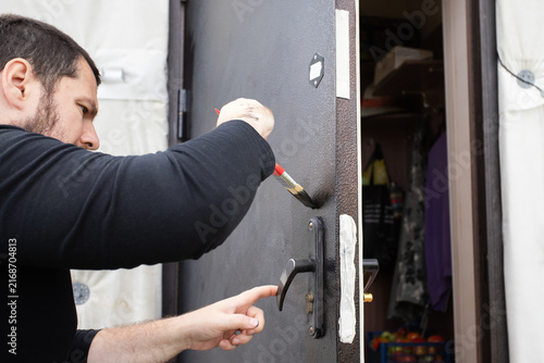 man carefully painting around the handle of an old steel entrance door, protecting masked hardware while restoring the exterior surface during diy home renovation