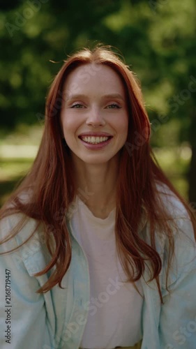 Vertical portrait of beautiful young woman with long red hair and radiant smile looking at camera. Cheerful Caucasian model with freckles enjoying sunny day outdoors in green summer park.