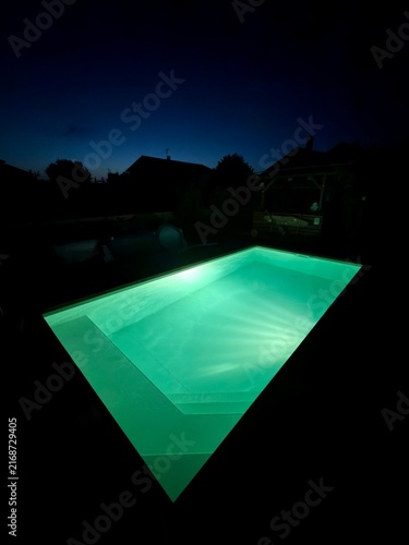 A green lit stepped swimming pool at night under a dark blue sky with houses in silhouette.