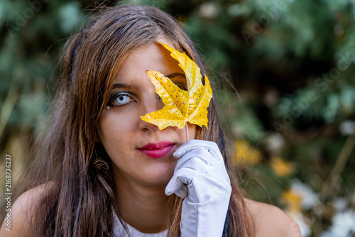 Artistic portrait of a woman with a yellow autumn leaf covering one eye, wearing a white glove and posing in a natural forest setting.