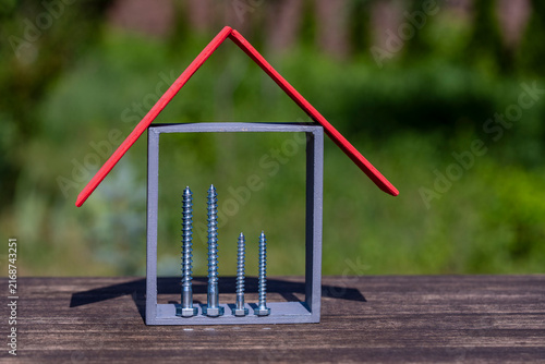 Tiny model house with red roof and silver screws inside on wooden surface against blurred green grass, closeup. Concept of house construction, building materials, mortgage or renovation cost