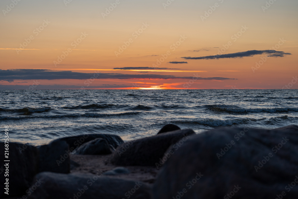 Obraz premium Baltic Sea sunset over gentle waves with dark coastal rocks in the foreground and a dramatic orange evening sky.