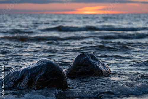 Canvas Print Wet coastal rocks surrounded by Baltic Sea waves at sunset, with a warm orange glow on the distant horizon