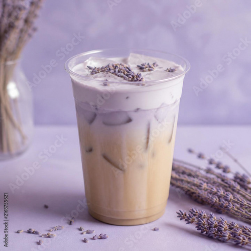 Iced lavender latte in a clear plastic cup with purple foam topping and dried lavender sprigs on a soft purple background