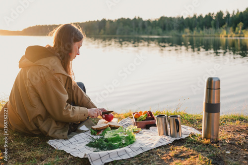 Woman preparing fresh sandwiches and vegetables during an outdoor picnic by a quiet lake, with a thermos and reusable mugs in warm evening sunlight