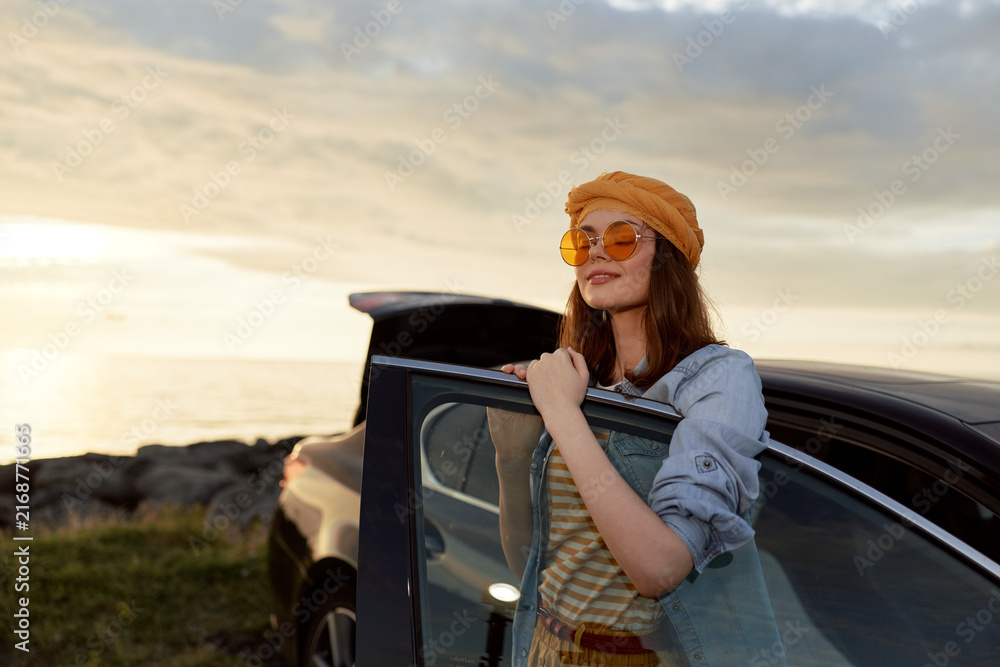 Fototapeta premium woman lifestyle fashion travel sunset outdoors car, Smiling woman by a car at sunset wearing denim jacket striped dress hat and sunglasses, an outdoor lifestyle moment