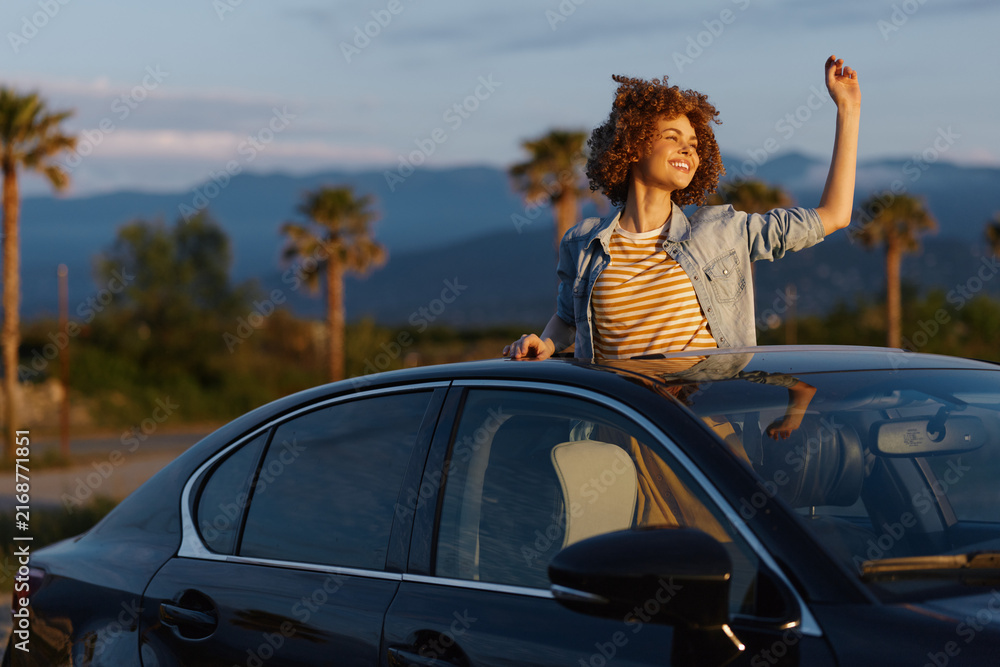 Fototapeta premium Joyful young woman leans out of a car window, raises her fist, as the sunset lights palm trees and distant mountains on a warm evening road trip.