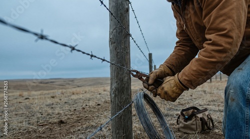 A close-up shot of a rancher wearing leather gloves and a brown jacket using pliers to repair a barbed wire fence on a wooden post in a dry, open pasture under an overcast sky.