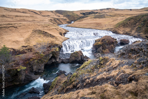 The Skogaa River flows through tranquil grass fields before forming the famous Skogafoss waterfall on a cloudy day in March.
