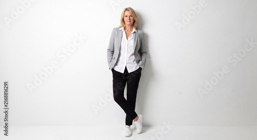 Mature woman standing in a grey blazer, white shirt, black trousers, and white sneakers
