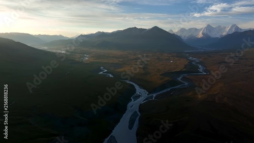 Aerial view of Sary Jaz river winding through the vast mountain valley at sunset with snow-capped peaks in the distance in Karakol, Issyk-Kul Region, Kyrgyzstan.