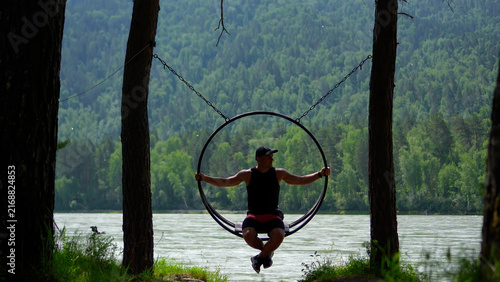 Cuadro en lienzo Active mid adult man in black sportswear relaxing and swinging in a round metal ring swing surrounded by dense Siberian pine forests