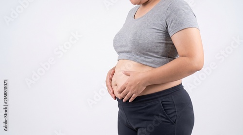 An overweight adult woman shown in side profile gently holding her abdominal area to highlight visible belly fat and excess weight, wearing simple neutral athletic clothing, isolated against a plain 