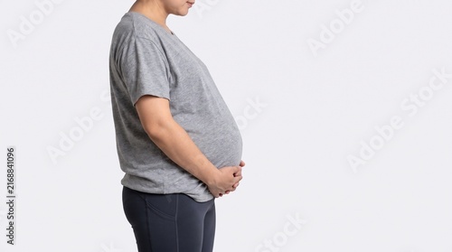 An overweight adult woman shown in side profile gently holding her abdominal area to highlight visible belly fat and excess weight, wearing simple neutral athletic clothing, isolated against a plain 
