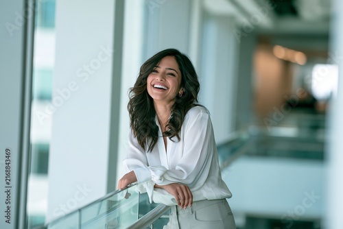 Happy young woman with curly hair laughing while leaning on a railing in a bright office space
