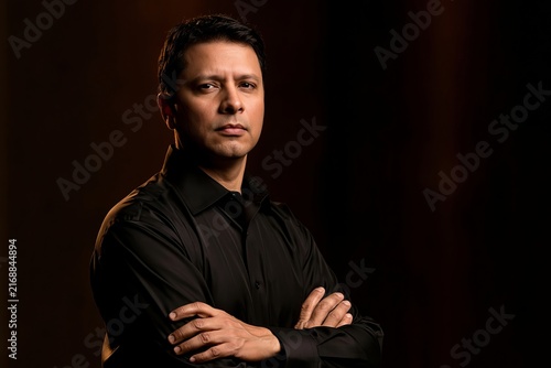Serious man with dark hair wearing a black shirt standing with his arms crossed in studio