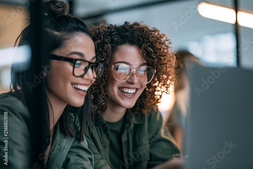 Two happy female colleagues wearing glasses smiling while looking at a laptop screen