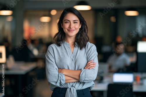Confident smiling businesswoman with crossed arms standing in a busy modern open plan office