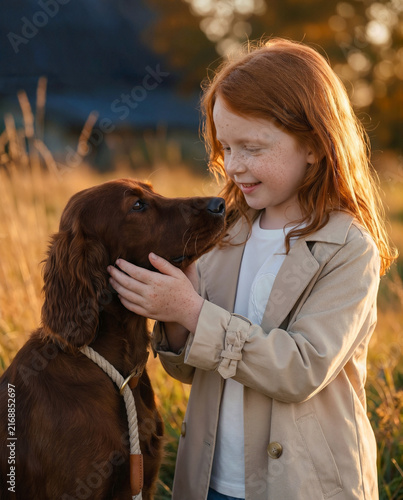 Rustic early autumn photoshoot of a little red-haired girl with a red setter dog near an old barn, warm countryside mood with soft seasonal light