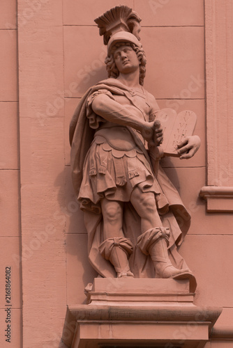 Bild auf Leinwand Eindrucksvolle antike Kriegerstatue mit Helm, Umhang und Gesetzestafeln an einer historischen Fassade