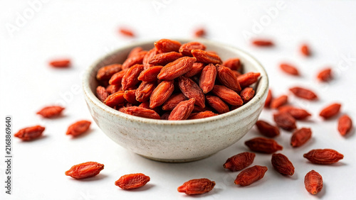 A white ceramic bowl filled with bright red dried goji berries, with several pieces scattered around the base on a clean white studio background.