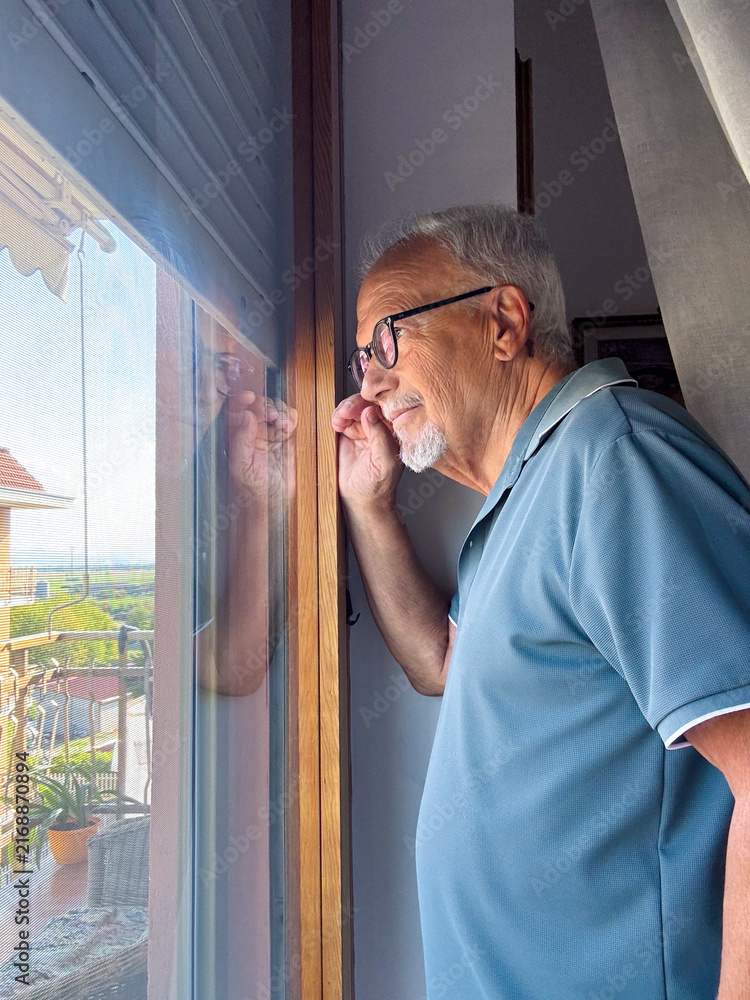 Fototapeta premium Elderly man looking through a window with reflection, thoughtful and contemplative at home on a balcony view
