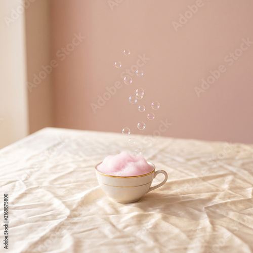Minimalist conceptual still life of a porcelain cup with pink foam and floating bubbles on wrinkled cream silk tablecloth