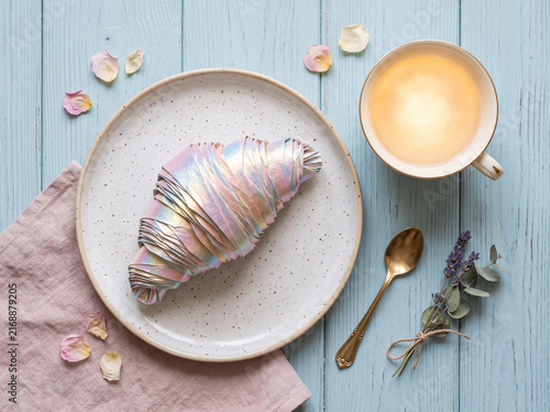 Aesthetic flat lay still life: iridescent holographic croissant on ceramic plate with cup of tea on pastel blue wooden table