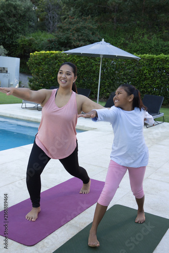 Diverse mother and daughter practicing standing yoga on tiled pool deck with purple and green mats