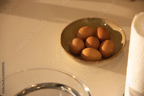 Ceramic bowl is sitting on countertop holding six brown eggs with blurred glass bowl, towel roll