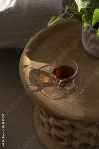 Clear glass teacup is resting on carved wooden side table near bed, sunlight casting amber glow