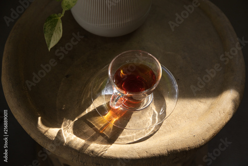 Clear glass teacup on saucer is sitting on textured table as sunlight is casting refracted patterns