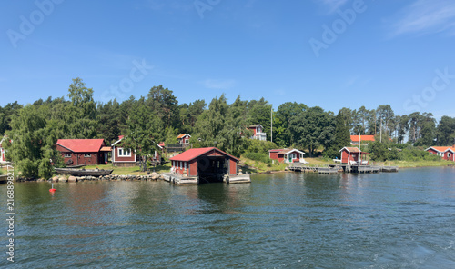 Konstfotografi Traditional red wooden houses and boathouses along the waterfront in the Stockholm Archipelago, Sweden, on a sunny summer day