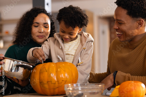 African American family scooping guts from carved pumpkin at kitchen counter with spoon and pot