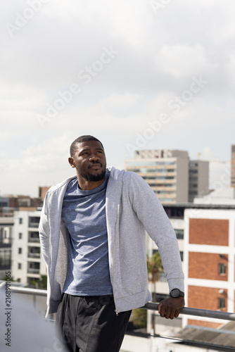 African American man leaning on rooftop metal railing, wearing gray zipup hoodie and watch