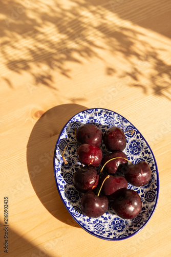 overhead shot of deep red cherries on a Portugese handmade blue and white dish on a light wood table with dappled shadows