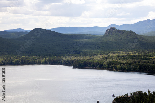 Scenic lake landscape with calm rippling water, lush green shoreline, resort buildings and gazebos surrounded by trees, with mountains visible in the distance under a dramatic cloudy sky. Peaceful out