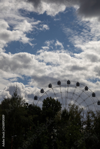 Ferris wheel silhouetted against a dramatic blue sky filled with large white clouds, viewed above a lush green park. Atmospheric outdoor amusement park scene with strong contrast between the dark tree